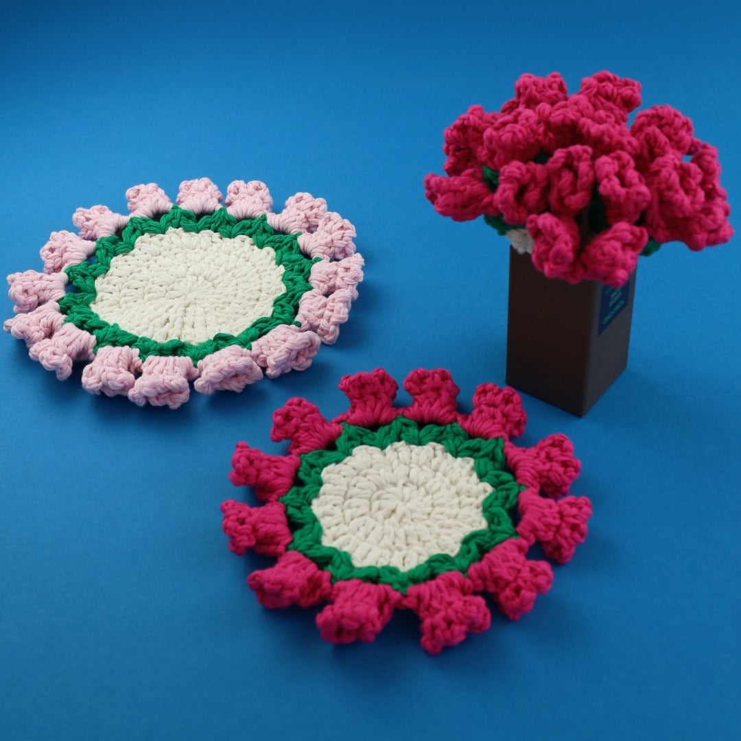 Two crocheted coasters with a central white circle, pink and green outer border, and a vase with pink flowers on a blue background.